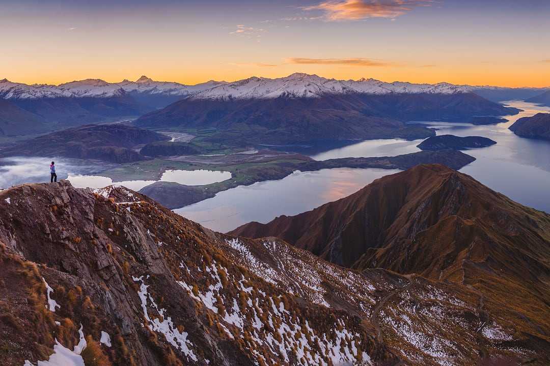 Hiker standing on a mountain trail, enjoying the beautiful sunrise over Lake Wanaka, New Zealand in winter