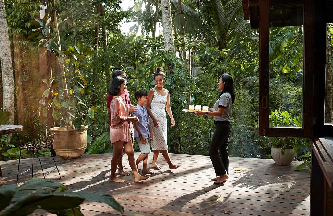 Bali, Indonesia A female specialist stands on a sunny tropical balcony, smiling and gesturing while speaking to a traveling family.