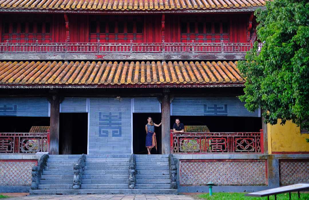 Travelers at the Imperial City of Hue, Vietnam, standing outside a historic temple with red columns and tiled roof.