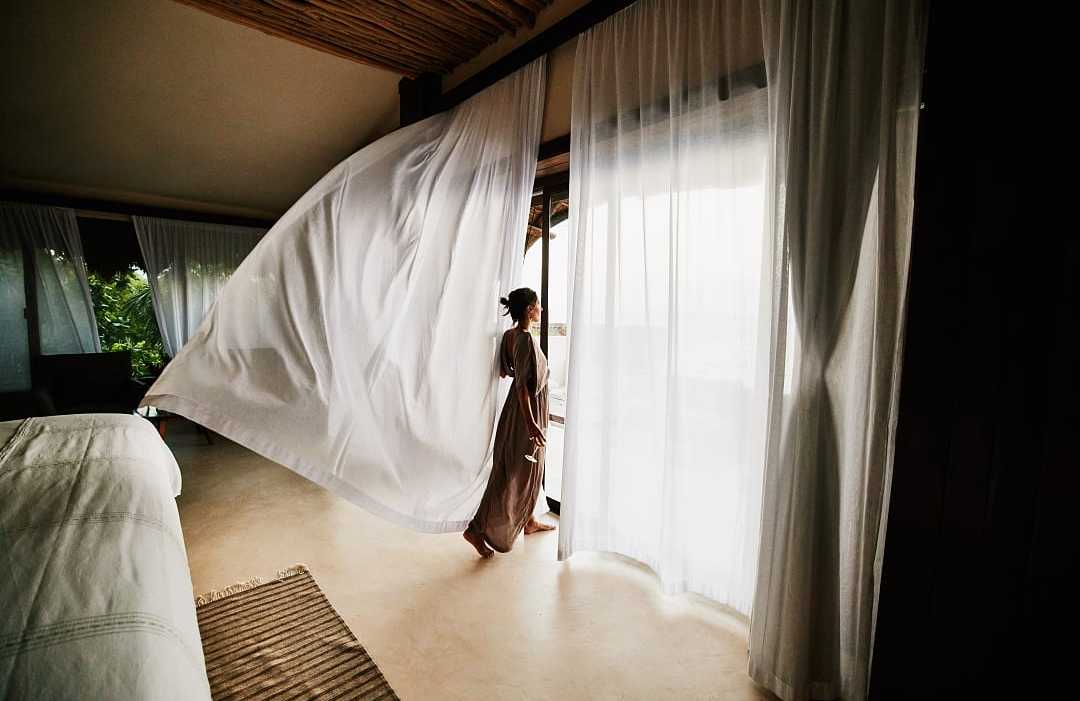 Luxury room in Mexico A woman in a flowing dress stands by large windows as a sheer white curtain billows in a luxury hotel room.