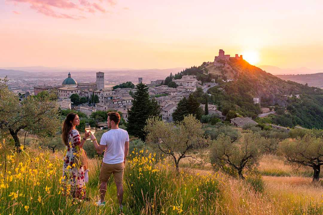 Couple toasting with wine at sunset overlooking a picturesque medieval town surrounded by olive trees and rolling hills