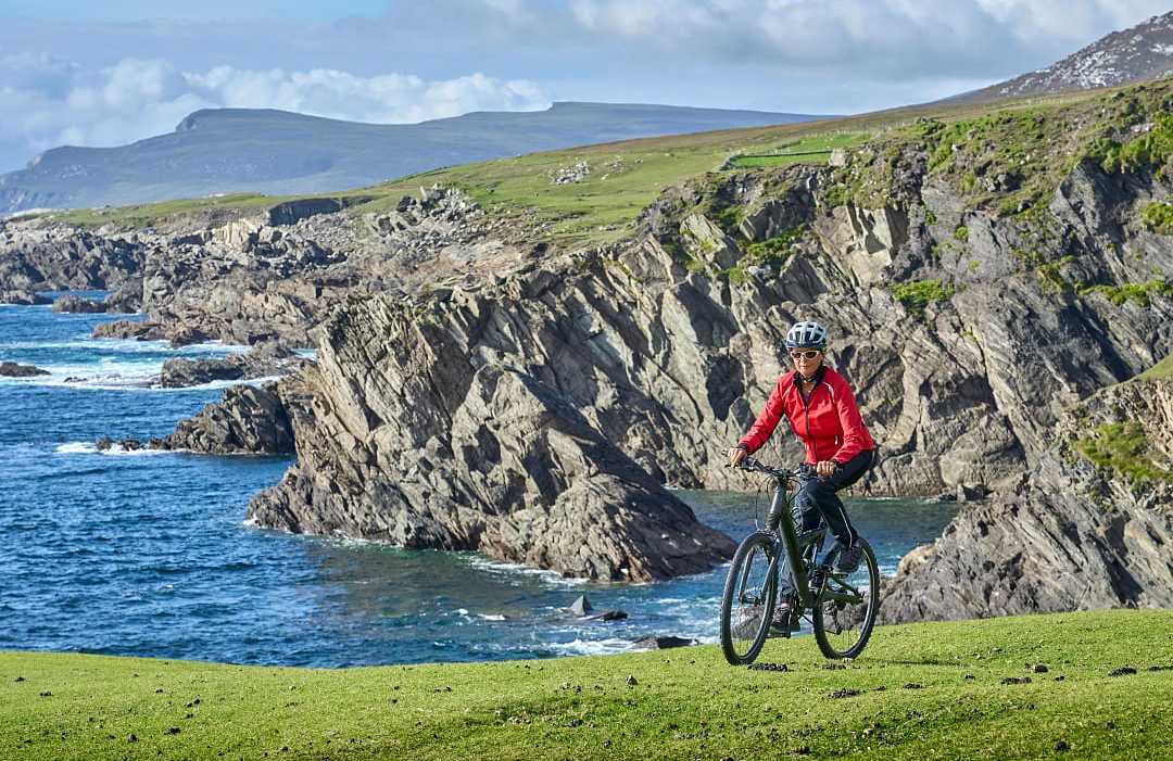 Senior cyclist biking along scenic Irish coastal cliffs with blue ocean waves and rugged landscapes