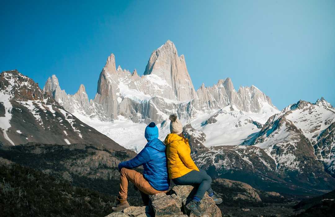 wo hikers in colorful jackets admire snowy Mount Fitz Roy in Patagonia under a clear blue sky