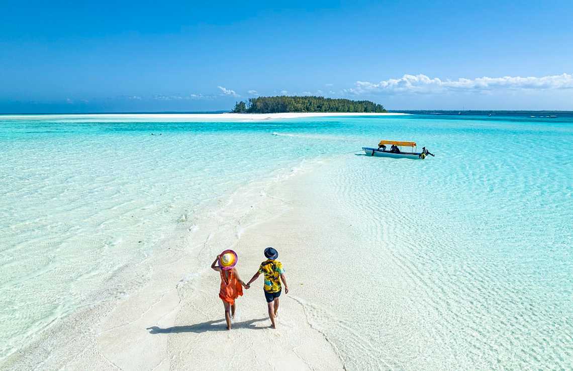 Couple walks on sandbank in clear turquoise water toward island in Zanzibar, Tanzania.