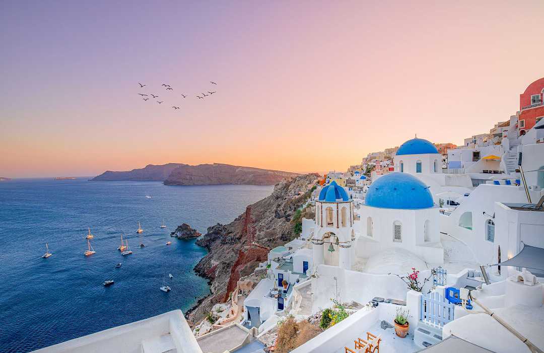 The sun sets over Oia’s blue-domed churches and Aegean Sea.