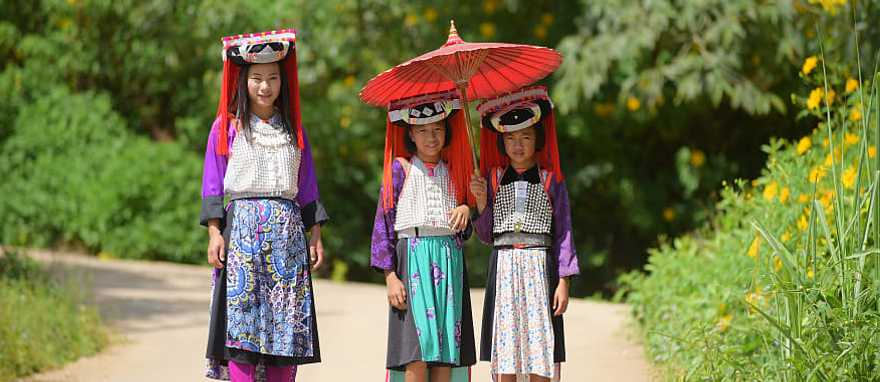 Three young Lahu Tribe girls in custom dress, smiling among the sunlight in Thailand.