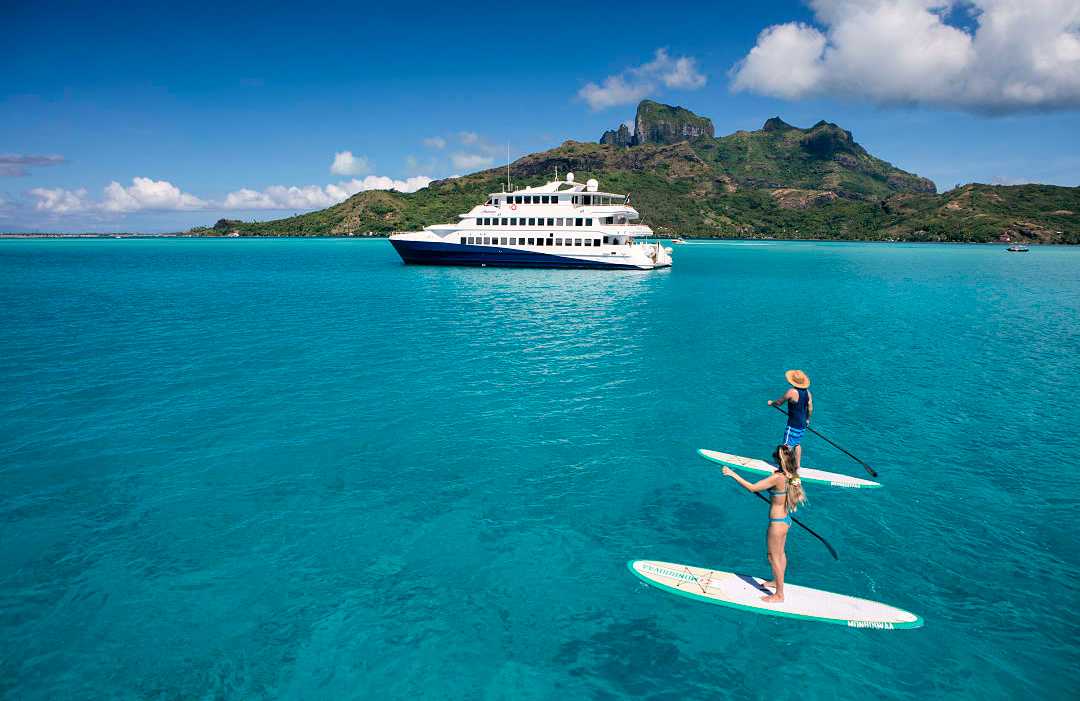 Bora Bora in French Polynesia.  Photo courtesy of Grégoire Le Bacon / Tahiti Tourism Couple paddleboarding on turquoise water in Bora Bora with a luxury yacht and tropical island backdrop