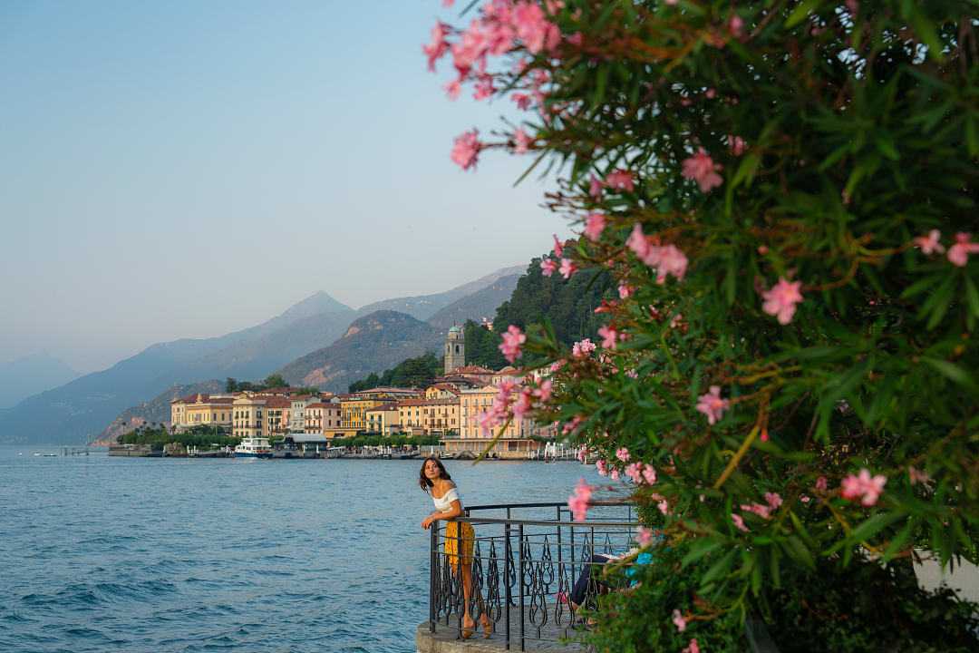 Scenic view of a woman by Lake Como, vibrant flowers, waterfront architecture, and mountains in the distance